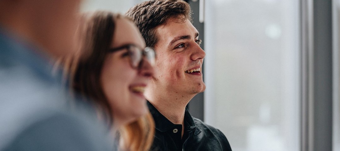 Zwei junge duale Studierende lächeln, während sie in einem lichtdurchfluteten Raum an einem Gespräch teilnehmen.