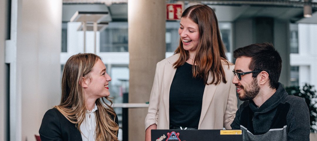 Lachende junge Absolventen in einer Teamdiskussion, während sie gemeinsam an dem Laptop vor ihnen arbeiten.