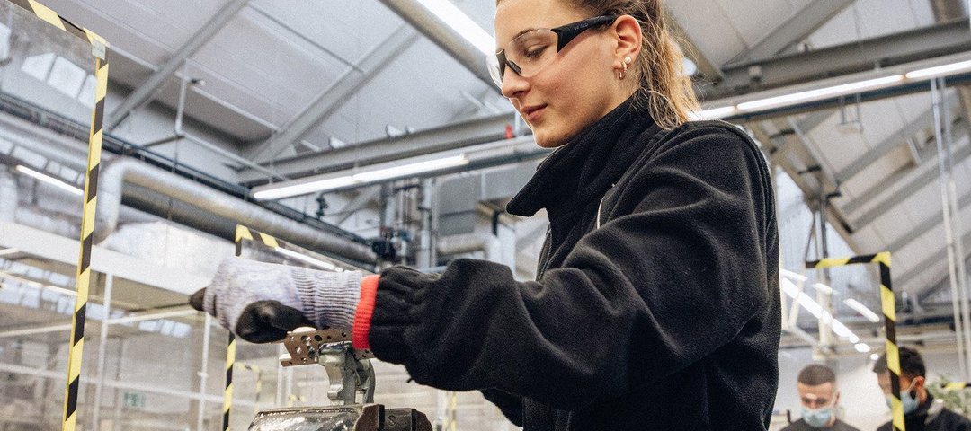 Eine technische Auszubildende mit Schutzbrille und Sicherheitshandschuhen arbeitet an einem Schraubstock in einer Werkstattumgebung.