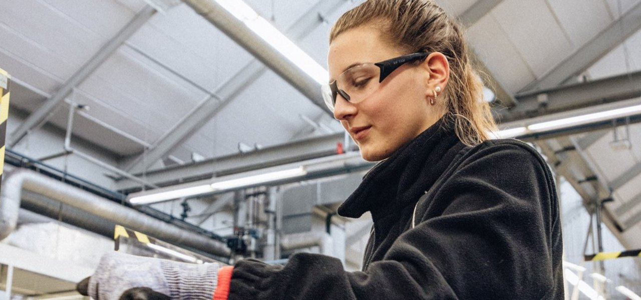 Eine technische Auszubildende mit Schutzbrille und Sicherheitshandschuhen arbeitet an einem Schraubstock in einer Werkstattumgebung.