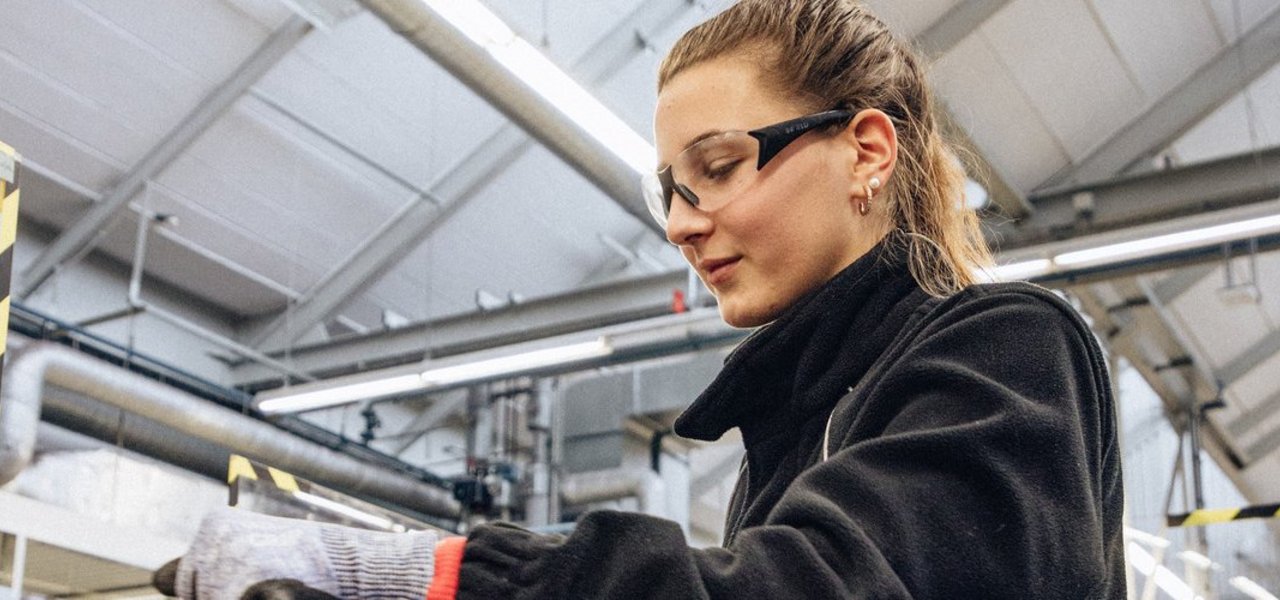Eine technische Auszubildende mit Schutzbrille und Sicherheitshandschuhen arbeitet an einem Schraubstock in einer Werkstattumgebung.