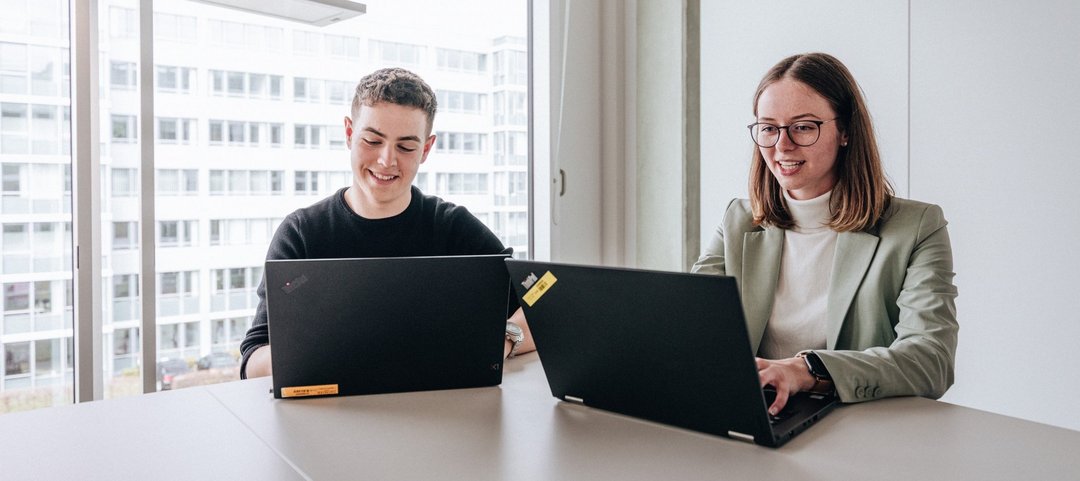 Zwei Doktoranten, ein junger Mann und eine junge Frau, sitzen nebeneinander und arbeiten am Laptop im Daimler Truck Campus.