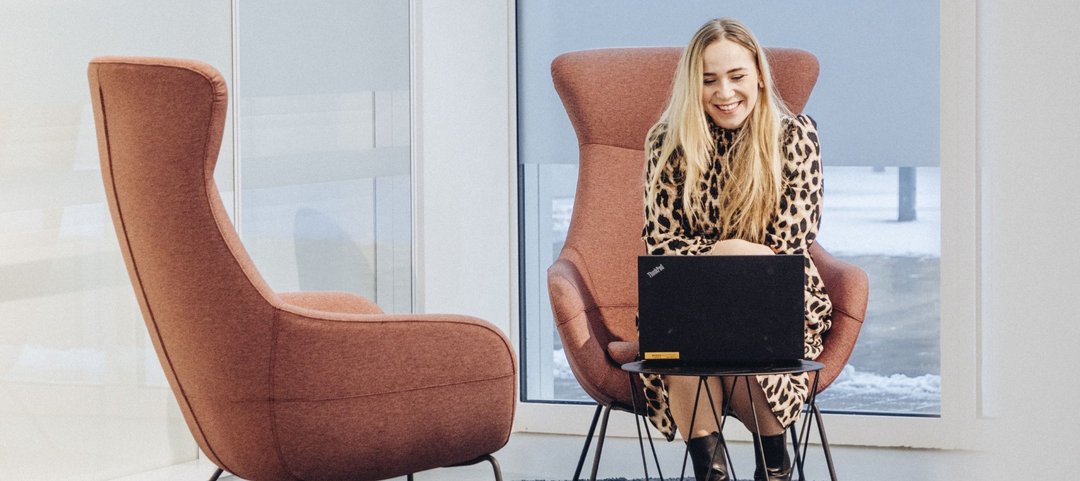 Eine junge Mitarbeiterin sitzt in einem modernen hellen Lounge-Bereich auf einem Sessel und arbeitet an einem Laptop, der auf einem kleinen Tisch vor ihr steht.